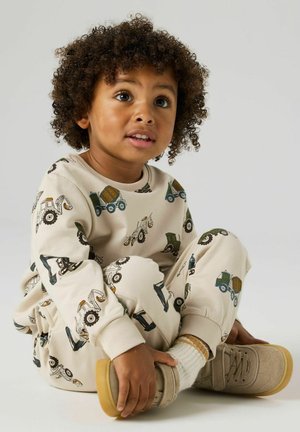 Toddler with curly hair sitting cross-legged, wearing cream-colored outfit with construction vehicle prints and beige shoes with white socks.