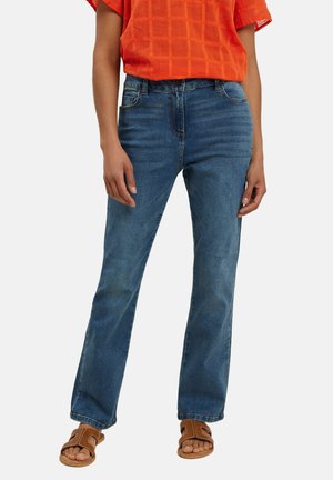 Kvinde iført blå jeans med rette ben, en kortærmet orange top og brune åbne sandaler, stående mod en ensfarvet baggrund.