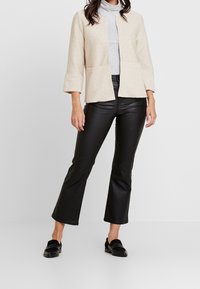 Svetle béžový texturovaný blazer s třičtvrtinovými rukávy, kombinovaný s černými rozšířenými kalhotami a černými mokasíny. Oblečení doplňuje jednoduchý šedý rolák.