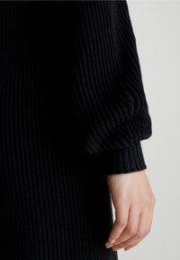 Robe en tricot noir à côtes avec des manches volantes et un poignet ajusté. Le tissu a une apparence texturée et une épaisseur moyenne.