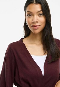Cardigan bordeaux a scollo a V in morbido tessuto a maglia, caratterizzato da punti aperti e una vestibilità rilassata. Indossato sopra una camicia bianca, senza motivi visibili.