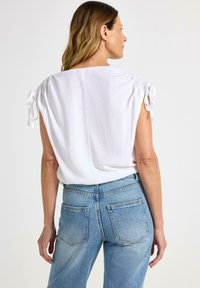 Hvid kortærmet bluse med samlede skulderdetaljer og bindebånd, parret med medium-vaskede denimjeans med baglommer.