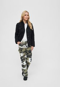 Musta kevyt takki, valkoinen t-paita ja vihreät camo-vaatteen taskuhousut. Saappaat viimeistelevät asun yksinkertaista taustaa vasten.