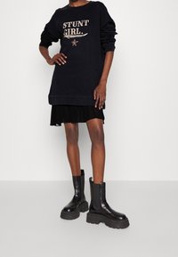 Svart sweatshirtklänning med texten "STUNT GIRL", kombinerad med en plisserad svart kjol och kraftiga svarta ankelskor med tjock sula.