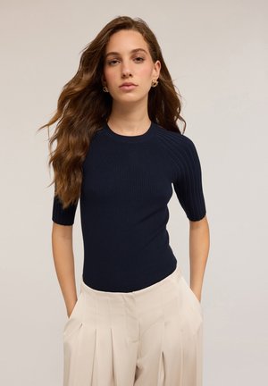 Top de punto acanalado en color marino con mangas cortas y cuello redondeado, combinado con pantalones plisados en color crema. Textura suave, diseño ajustado.