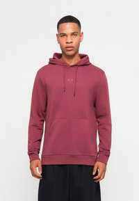 Burgunderfarget hettegenser med frontlomme, snor og en liten logo på brystet. Myk tekstur og avslappet passform, kombinert med mørke shorts.