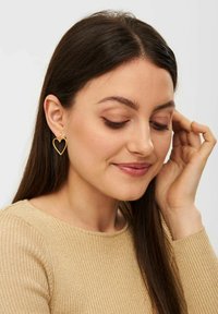 Orecchini a forma di cuore in oro appesi all'orecchio. Il design è minimalista con una finitura lucida, arricchito da piccoli accenti di perle.