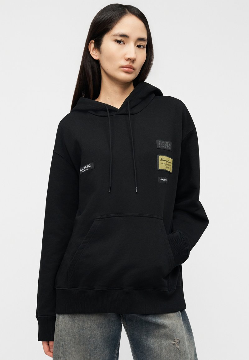 Black hooded sweatshirt with front pocket, featuring multiple fabric patches in varying colors and textures. Casual fit, ribbed cuffs.
