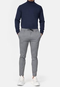 Marineblauer Rollkragenpullover aus Strick. Hellgraue, zulaufende Hose mit Kordelzug und Seitentaschen. Weiße Freizeitsneaker.