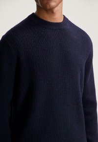 Man in een donker marineblauwe geribbelde gebreide trui met een ronde hals tegen een effen lichte achtergrond.