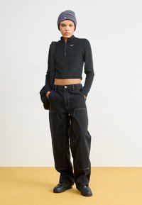 Czarna krótka bluza na zamek z długimi rękawami i małym logo, w połączeniu z luźnymi czarnymi dżinsami z przednimi kieszeniami i widocznym przeszyciem.