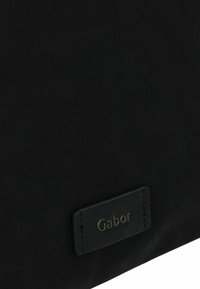 Sac à main noir en matériau lisse, présenté avec une étiquette en cuir cousue arborant le logo "Gabor" en relief. Design minimaliste avec une marque discrète.