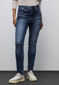 Slim fit jeans wykonane z ciemnoniebieskiego denimu; mają klasyczny pięciokieszeniowy design i subtelne wyblaknięcia. Noszone z jasnymi trampkami.
