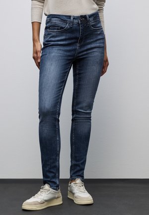 Jean skinny en denim bleu foncé ; présente un design classique à cinq poches et un léger délavage. Porté avec des baskets de couleur claire.