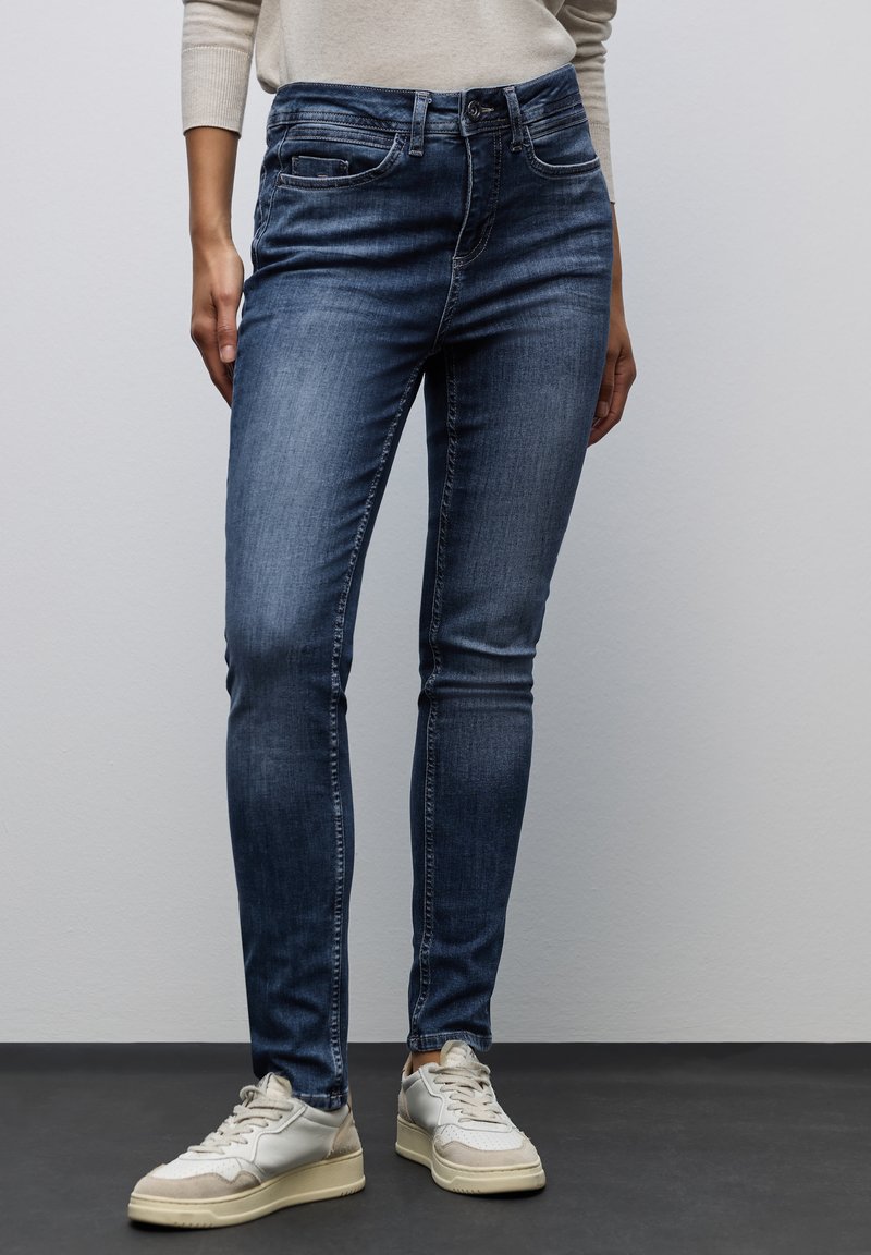 Jean skinny en denim bleu foncé ; présente un design classique à cinq poches et un léger délavage. Porté avec des baskets de couleur claire.