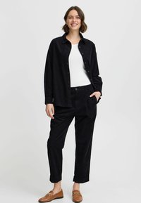 Outfit composto da una camicia e pantaloni di velluto a coste neri, con fit rilassato, chiusura con bottoni e orli dei pantaloni risvoltati. Indossato con mocassini marroni.