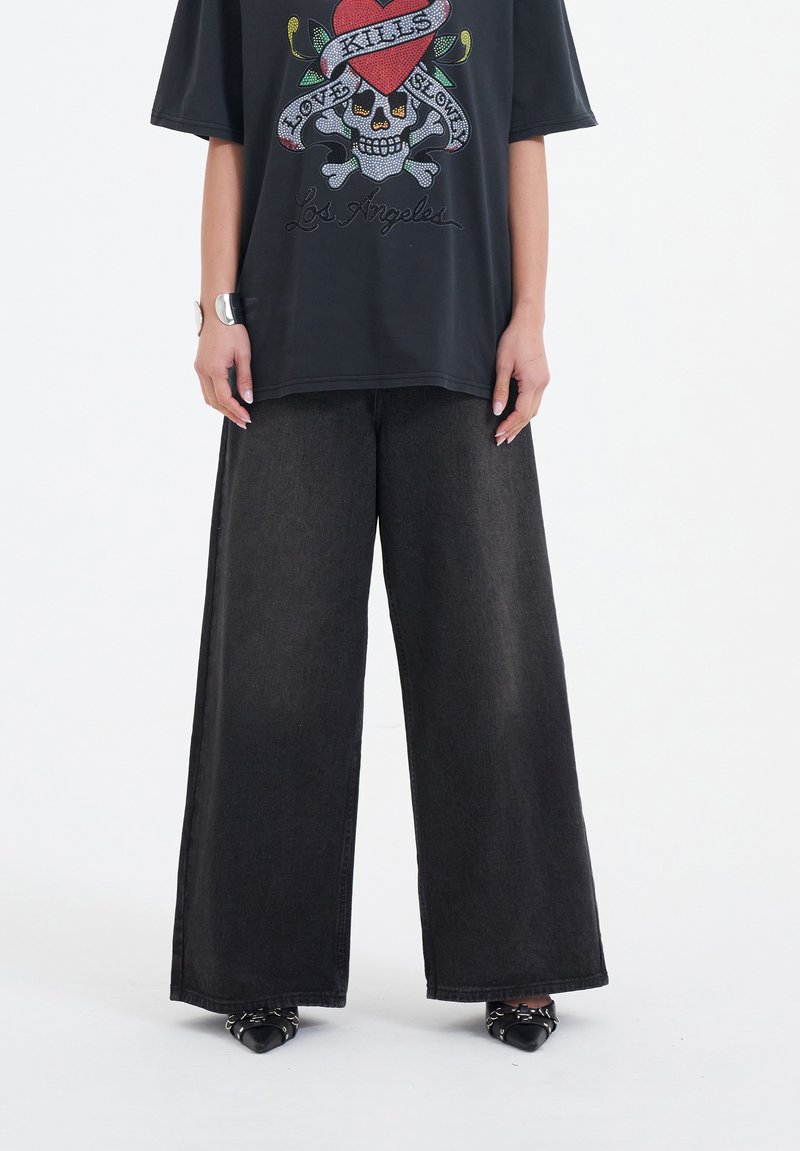 Schwarzes �übergroßes T-Shirt mit einem bunten Totenkopf-Graphic und Textdesign, kombiniert mit weiten schwarzen Jeanshosen und spitz zulaufenden schwarzen Schuhen.