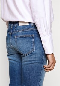 Jean en denim bleu avec une finition délavée, design à cinq poches et une étiquette blanche à l'arrière. Associé à une chemise boutonnée rose clair.