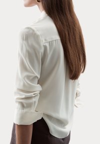 Blusa color crema con mangas largas, puños enrollados y una tela suave y ligera, que presenta un drapeado sutil y un cierre con botones en la parte posterior.