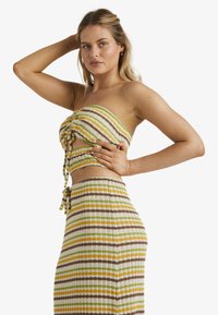 Frau in einem gestrickten, trägerlosen Crop-Top und einem passenden hochtaillierten Rock mit horizontalen Streifen in Grün, Gelb, Braun und Weiß.