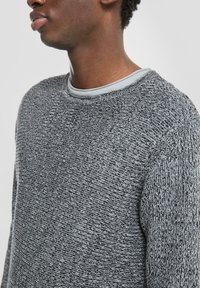 Grijze gebreide trui met een gemêleerd patroon, ronde halslijn en gelaagd met een lichtgrijze T-shirt eronder.