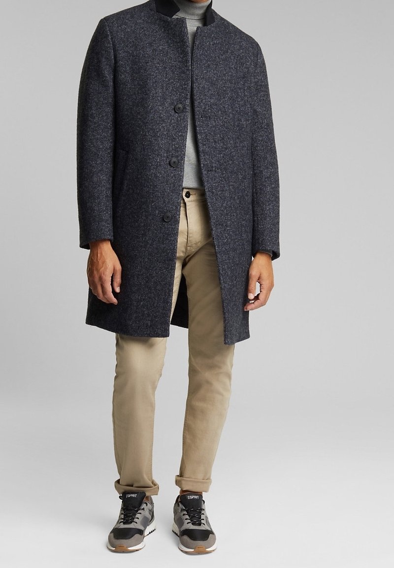 Uomo che indossa un cappotto di lana grigio scuro con bottoni sopra un dolcevita grigio chiaro, pantaloni beige e sneakers grigio-neri su uno sfondo grigio uniforme.