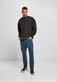 Hombre de pie con las manos en los bolsillos, vistiendo un sudadera negra, jeans azules y zapatillas de cuadros en blanco y negro, sobre un fondo gris liso.