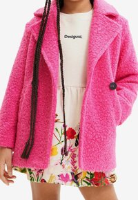 Manteau rose fuchsia texturé avec un col cranté et une fermeture à un bouton, porté sur une robe à motif floral avec une taille froncée.
