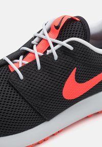 Sapatilha atlética em malha preta com sola cinza, apresentando detalhes e atacadores laranja. Inclui um destacado logótipo laranja da Nike na lateral.