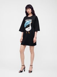 Jeune femme portant une robe noire oversize avec un motif d’une oie tenant un parapluie bleu et le texte Moschino, associée à des sandales noires à talons hauts.