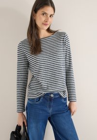 Haut à manches longues rayé bleu marine et blanc, en tissu texturé ; associé à un jean en denim bleu. Le mannequin tient un sac noir.