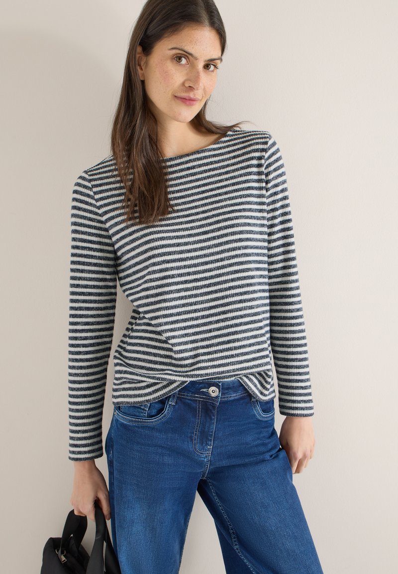 Haut à manches longues rayé bleu marine et blanc, en tissu texturé ; associé à un jean en denim bleu. Le mannequin tient un sac noir.