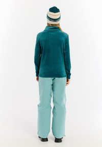Persoon draagt een teal gestructureerde jas, lichtblauwe broek, zwarte laarzen en een teal-en-witte beanie, staand met de rug naar de camera op een witte achtergrond.