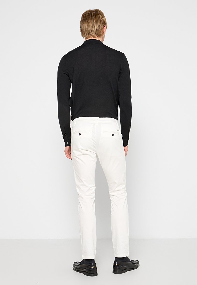 Camicia nera a maniche lunghe con un design aderente, abbinata a pantaloni bianchi slim-fit. I pantaloni presentano tasche posteriori con bottoni neri.