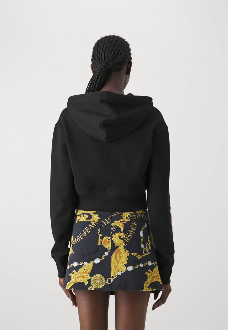 Haut de survêtement noir coupé avec capuche, associé à une jupe courte ornée d'un motif baroque doré et bleu. La jupe a des poches et des accents détaillés.