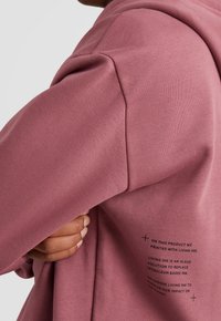 Sudadera rosa hecha de tejido suave. Presenta una capucha, mangas amplias y un texto impreso en el lado inferior izquierdo sobre tinta ecológica.