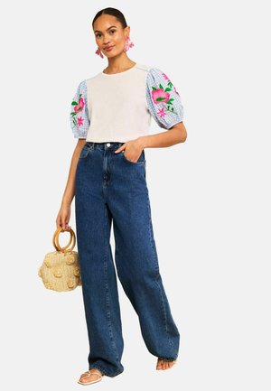 Woman wearing white top with puffed floral embroidered sleeves, wide-leg blue jeans, holding round wicker handbag, standing barefoot.