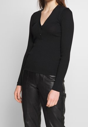 Top lungo a maniche ribbed nera con scollatura a V e tre bottoni sul davanti. Indossato con pantaloni in pelle nera, che mostrano un design aderente.