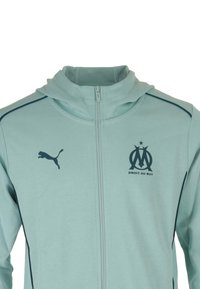 Giacca zip-up azzurro chiaro con cappuccio, caratterizzata da un logo scuro e dettagli sulle spalle e sul petto. Realizzata in tessuto morbido e testurizzato.