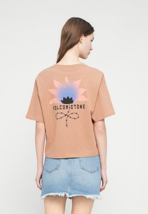 T-shirt en coton brun à manches courtes, avec un motif coloré de fleur et le texte "VOLCOM STONE" en noir. Associé à une jupe en denim.