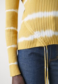 Żółty sweter prążkowany z białymi paskami, z dekoltem w kształcie kopertowym, długimi rękawami i wiązaniem na dole, zestawiony z ciemnymi dżinsami.