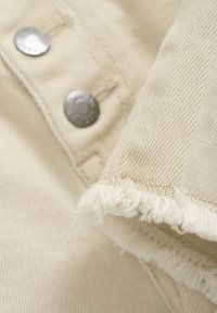 Beige tygdel med silverfärgade metallknappar, fransiga kanter och strukturerad väv, som framhäver släta och grova mönster.