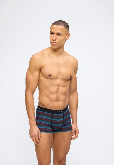 Μυώδης άνδρας όρθιος, φορώντας ριγέ μπλε και μαρουαν μποξεράκια, κοιτάζει προς τα αριστερά του πάνω σε απλό φωτεινό φόντο.