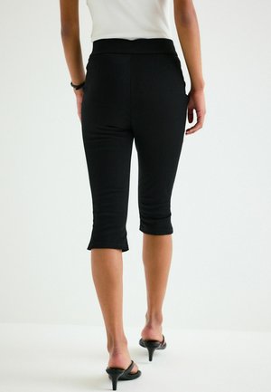 Femeie purtând pantaloni scurți negri, top alb și sandale negre cu toc, stând pe un fundal alb simplu.