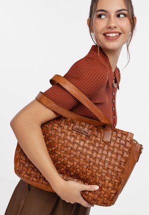 Femme portant un haut couleur rouille et tenant un grand sac à main Pikolinos en cuir marron tressé sur son épaule, souriante et regardant de côté.