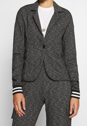 Personne portant un blazer texturé noir et blanc avec un pantalon assorti présentant des poignets rayés et une chemise blanche en dessous, accessoirisée d'un pendentif rond.