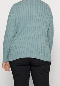 Ljusblå kabelstickad tröja med ribbad nederkant, långa ärmar och en avslappnad passform. Buren över svarta byxor som visar upp strukturell detaljering.