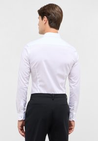 Camicia bianca con colletto strutturato, maniche lunghe e gemelli, abbinata a pantaloni tailored scuri che mostrano una finitura liscia.