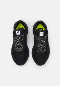 Des chaussures de sport noires avec un dessus texturé, des lacets blancs et noirs contrastants, et des semelles intérieures vert vif, arborant un logo sur la languette.