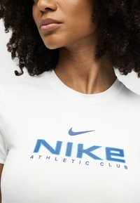 Γυναίκα που φοράει λευκό t-shirt Nike με μπλε λογότυπο και κείμενο "Athletic Club", κοντινό πλάνο στο λαιμό, το πηγούνι και το πάνω μέρος του στήθους.
